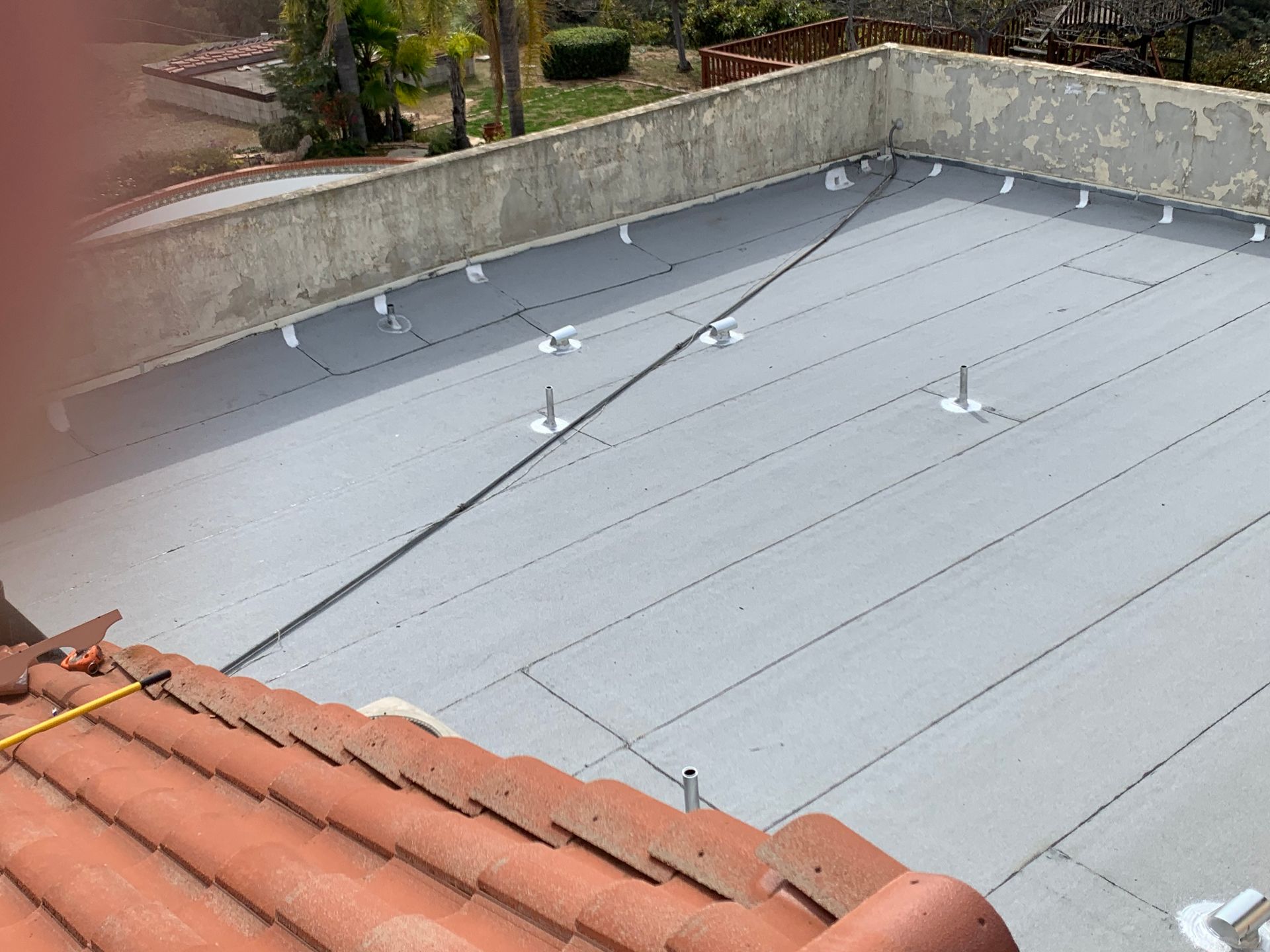 Flat rooftop with a gray membrane and rooftop vents beside terracotta roof tiles and a low concrete wall.