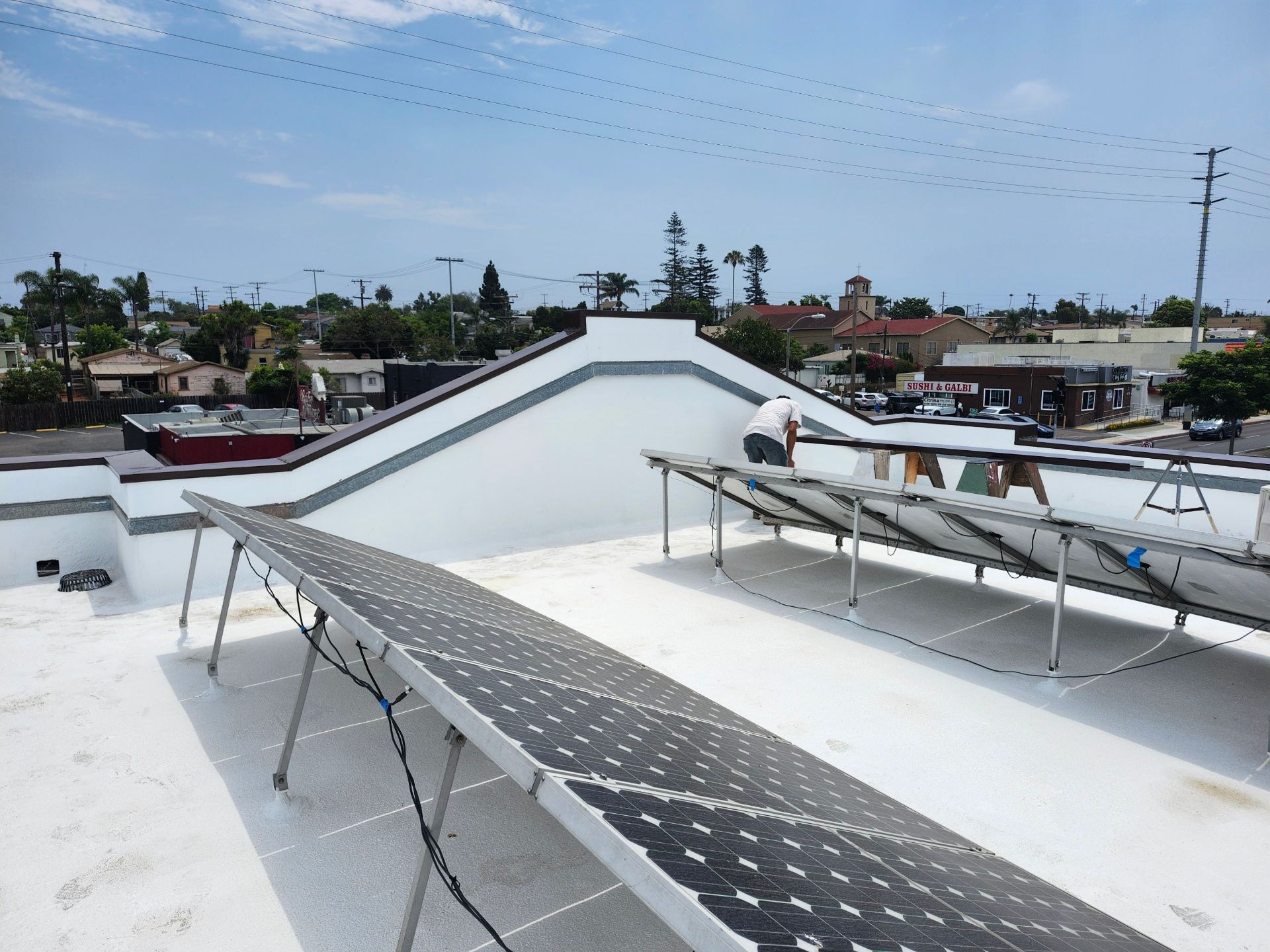 White rooftop with solar panels and piping under a blue sky, seen over a low urban skyline