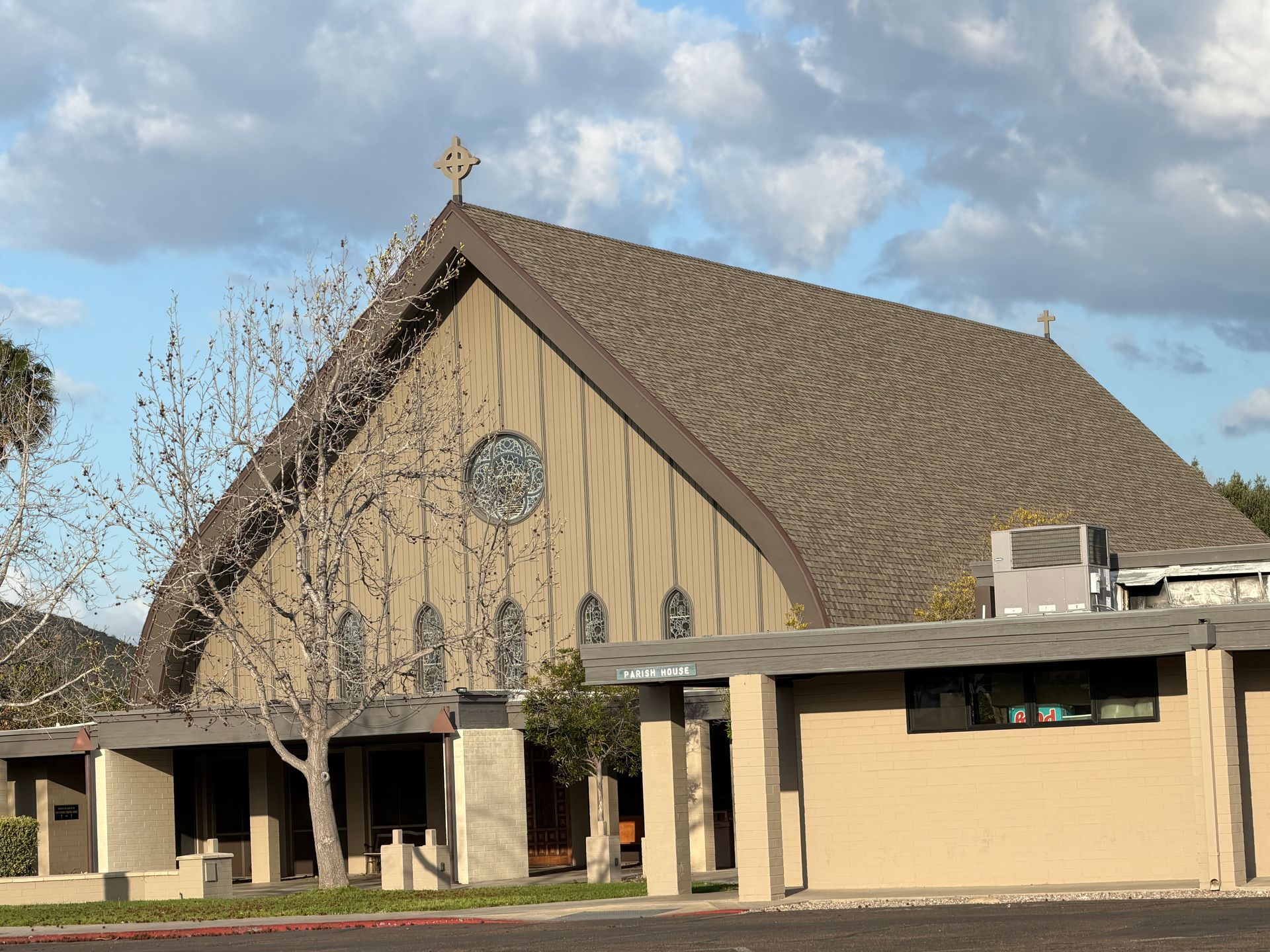 Modern church with tan exterior, steep roof, and stained-glass window under a cloudy sky