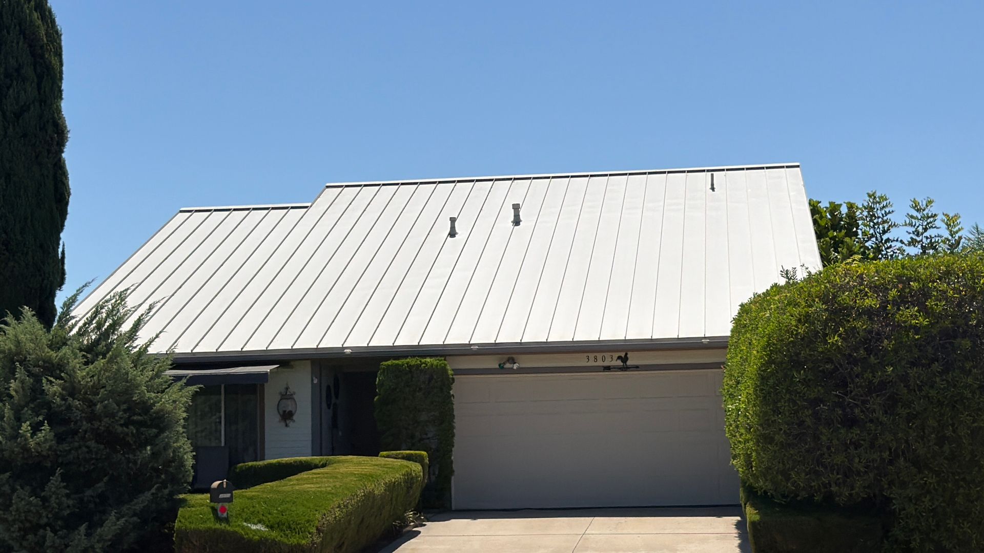 Single-story house with a white metal roof, garage, and trimmed green hedges under a clear blue sky