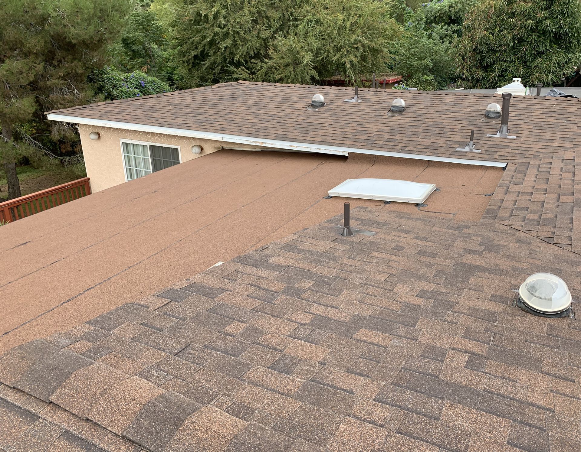 Roof with brown shingles, skylight, vent, and adjacent house exterior under trees
