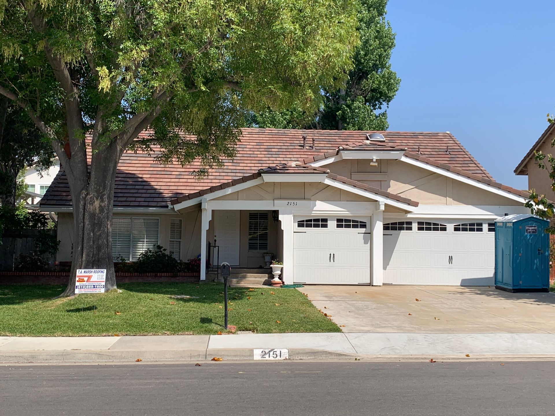 Suburban house with a two-car garage, large tree, and blue portable toilet by the driveway