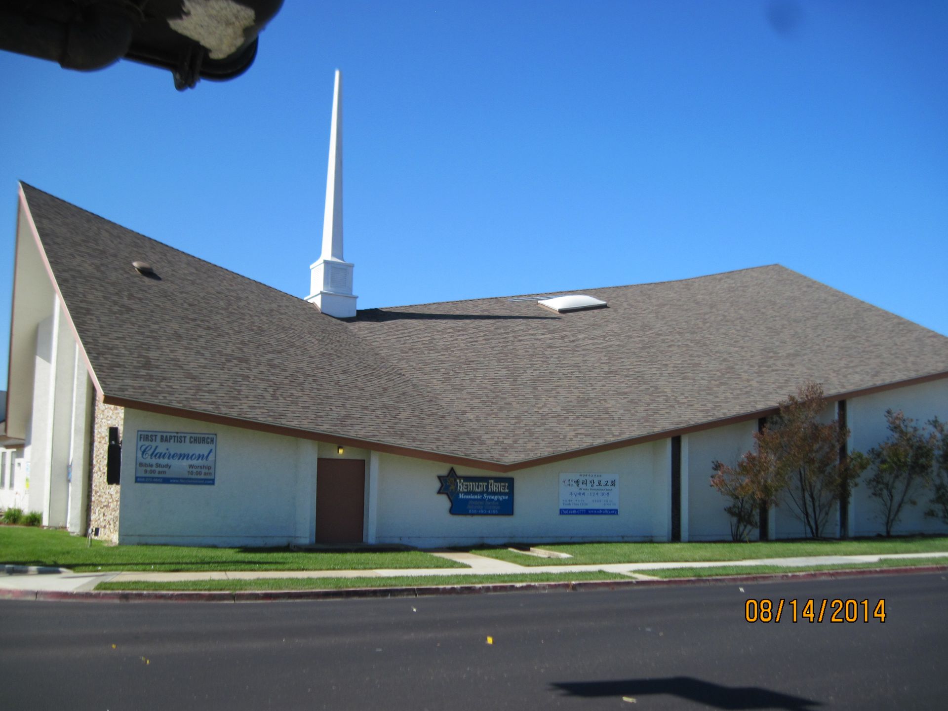 Small white church with a steep brown roof, blue sky, and a tall white steeple by the road.