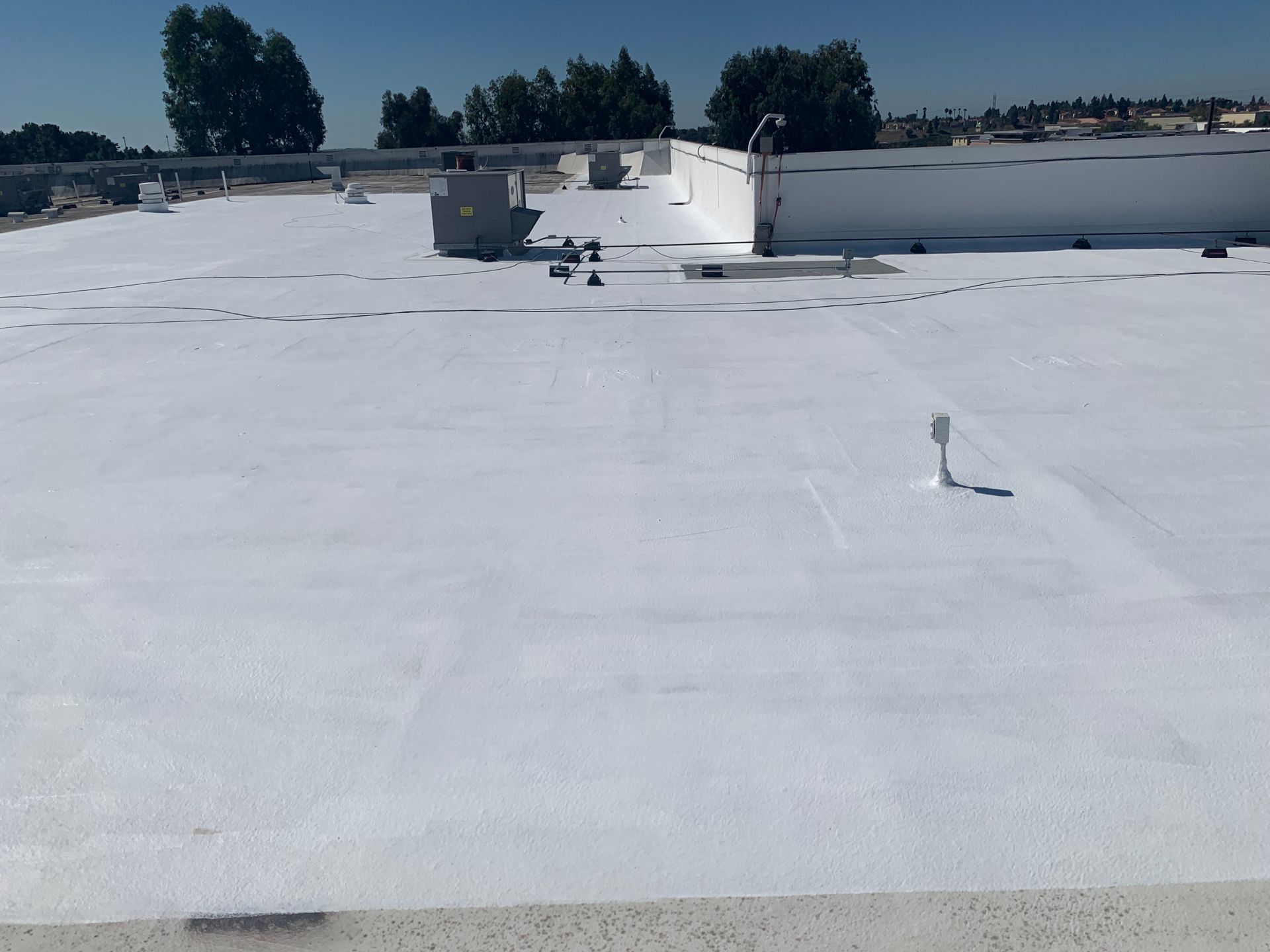 Flat white rooftop with vents and equipment under a clear sky, trees in the distance