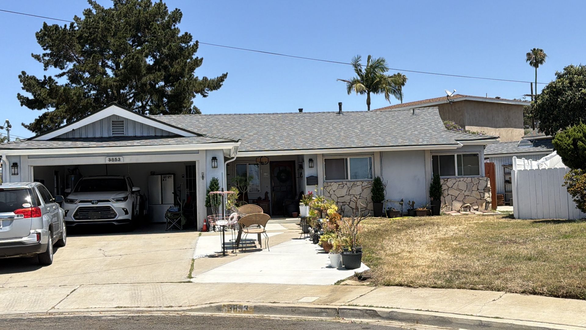 Single-story suburban house with two cars in the driveway and a dry front yard under a bright sky