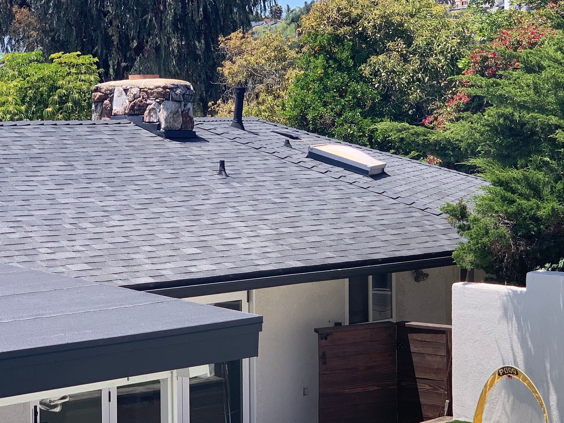 Roof with black shingles, skylight, chimney, and trees in the background.