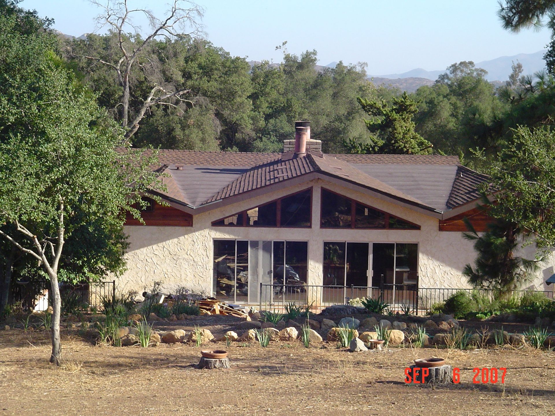 Single-story house with a brown roof and front windows, surrounded by trees and a rocky yard.