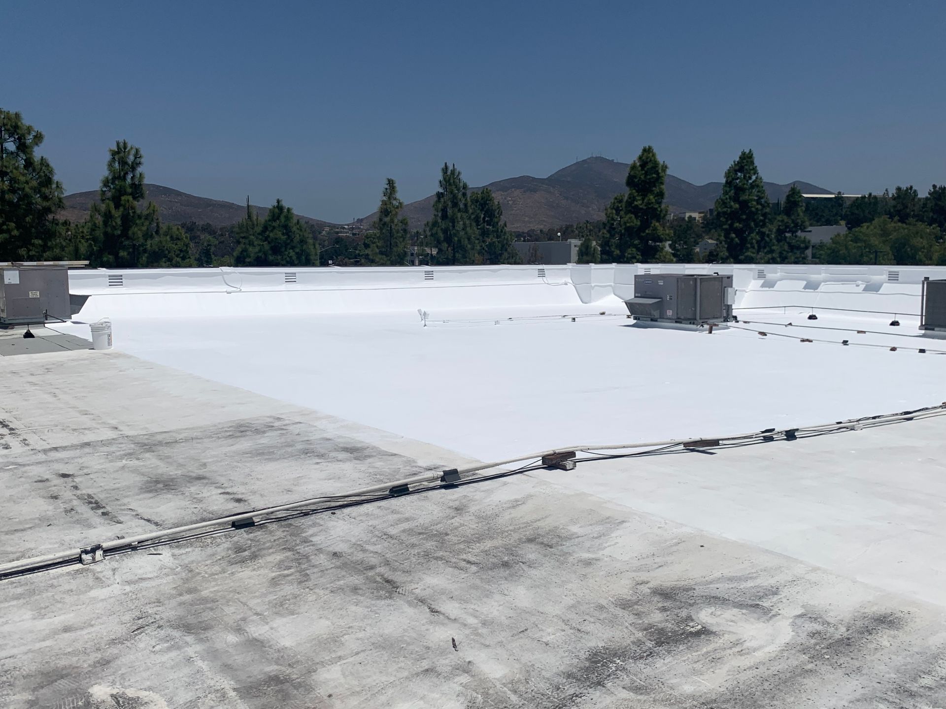 Flat white rooftop with vents, trees, and mountains under a clear blue sky.