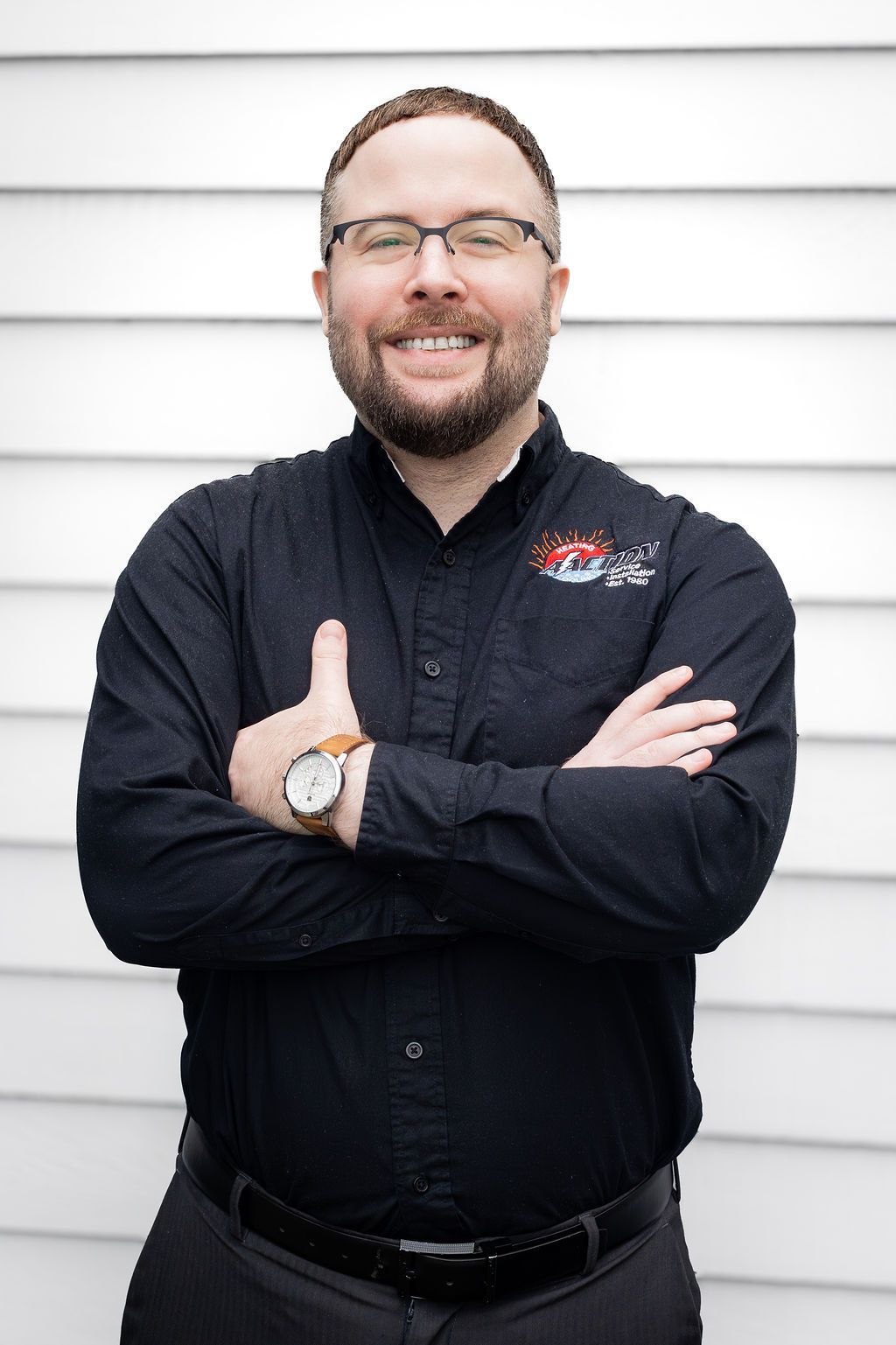 Smiling person in a black shirt with arms crossed, standing against white siding.
