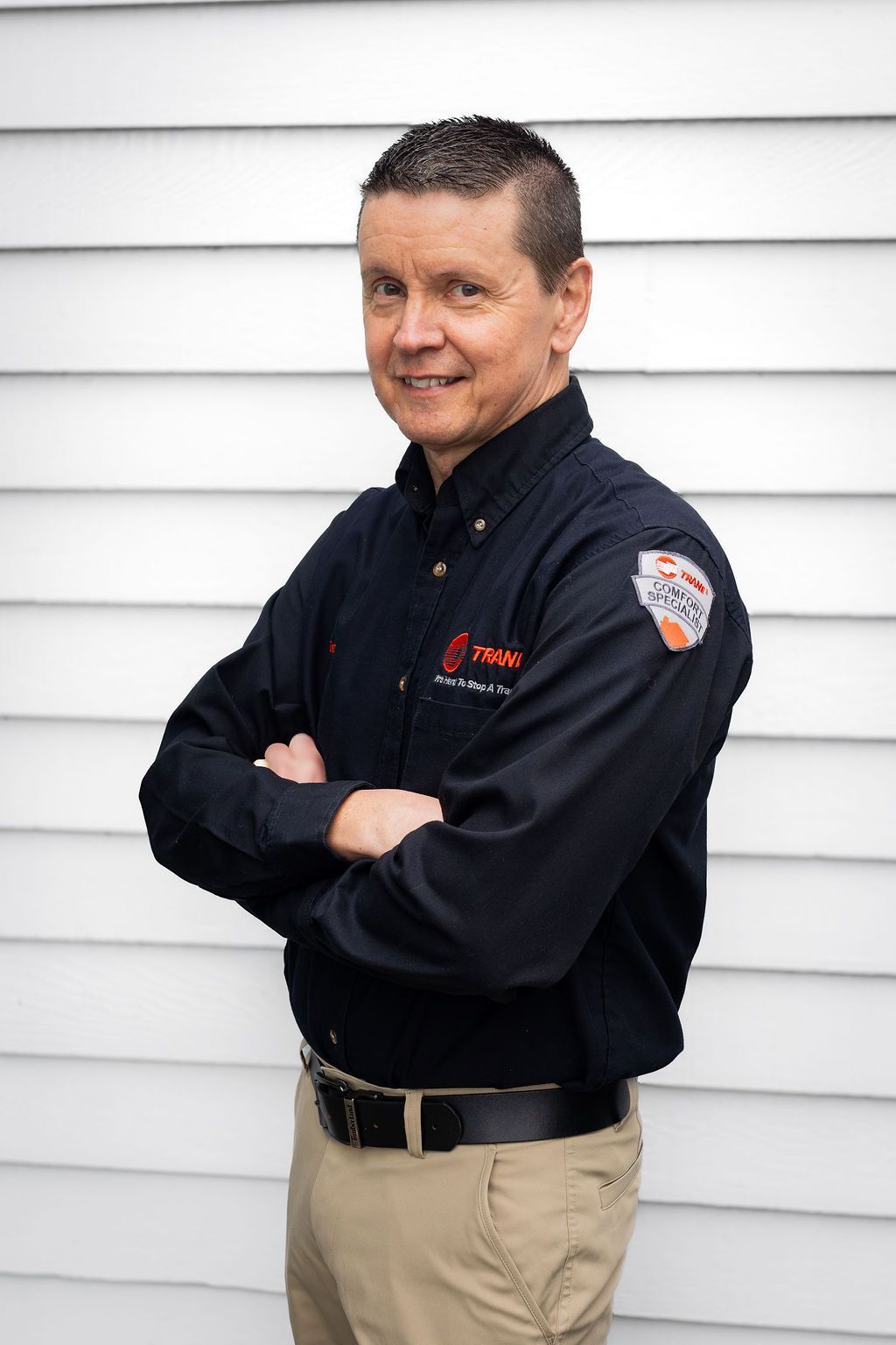 Person in a black uniform shirt and khaki pants stands with arms crossed against a white siding wall.