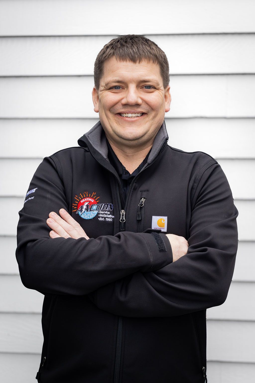 Smiling person in a black jacket standing with arms crossed against white siding.
