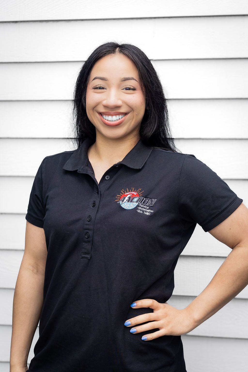 Woman in black polo shirt smiling with hand on hip against a white siding wall.
