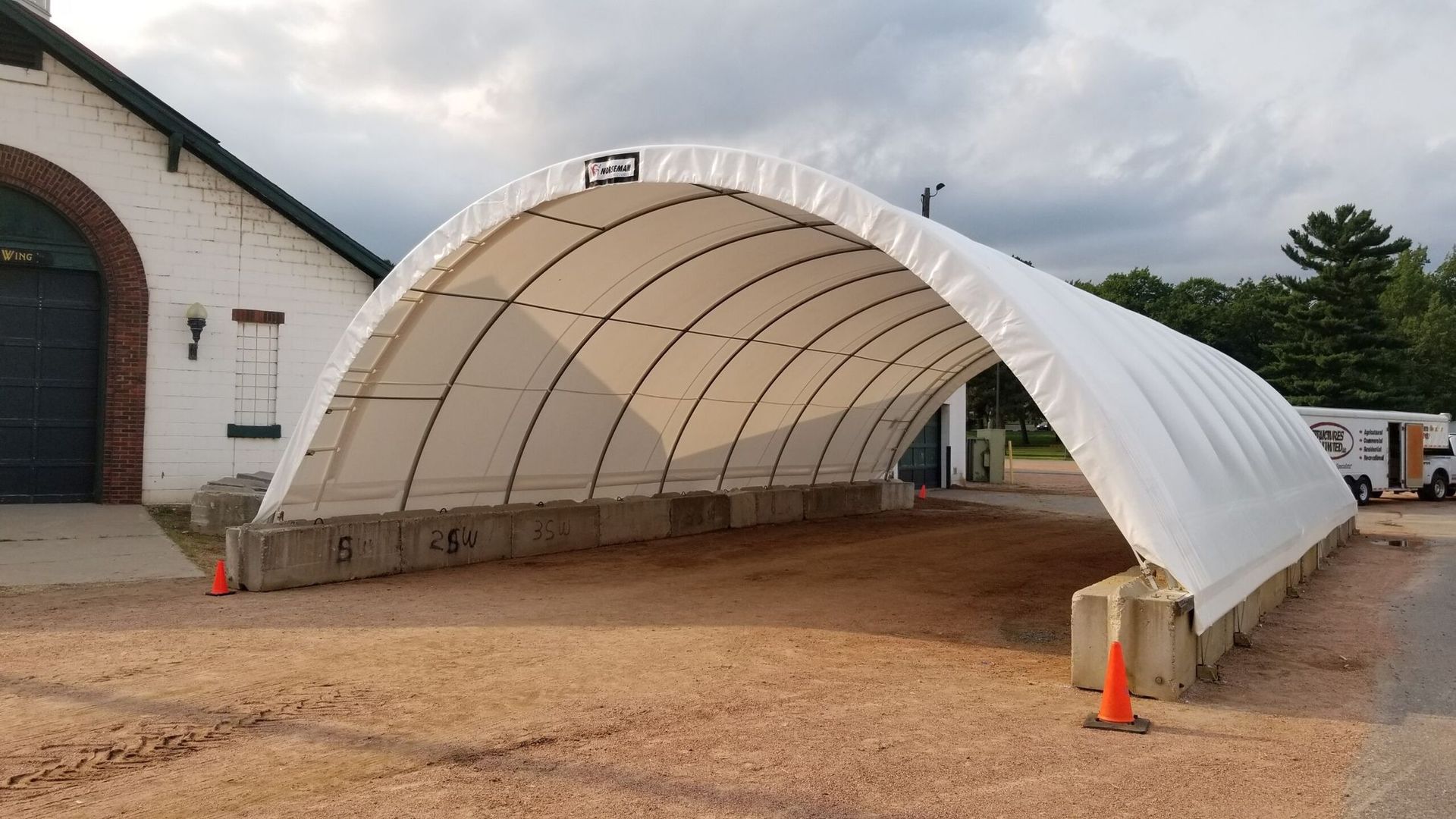 White arched shelter with concrete base; a building is on the left side.