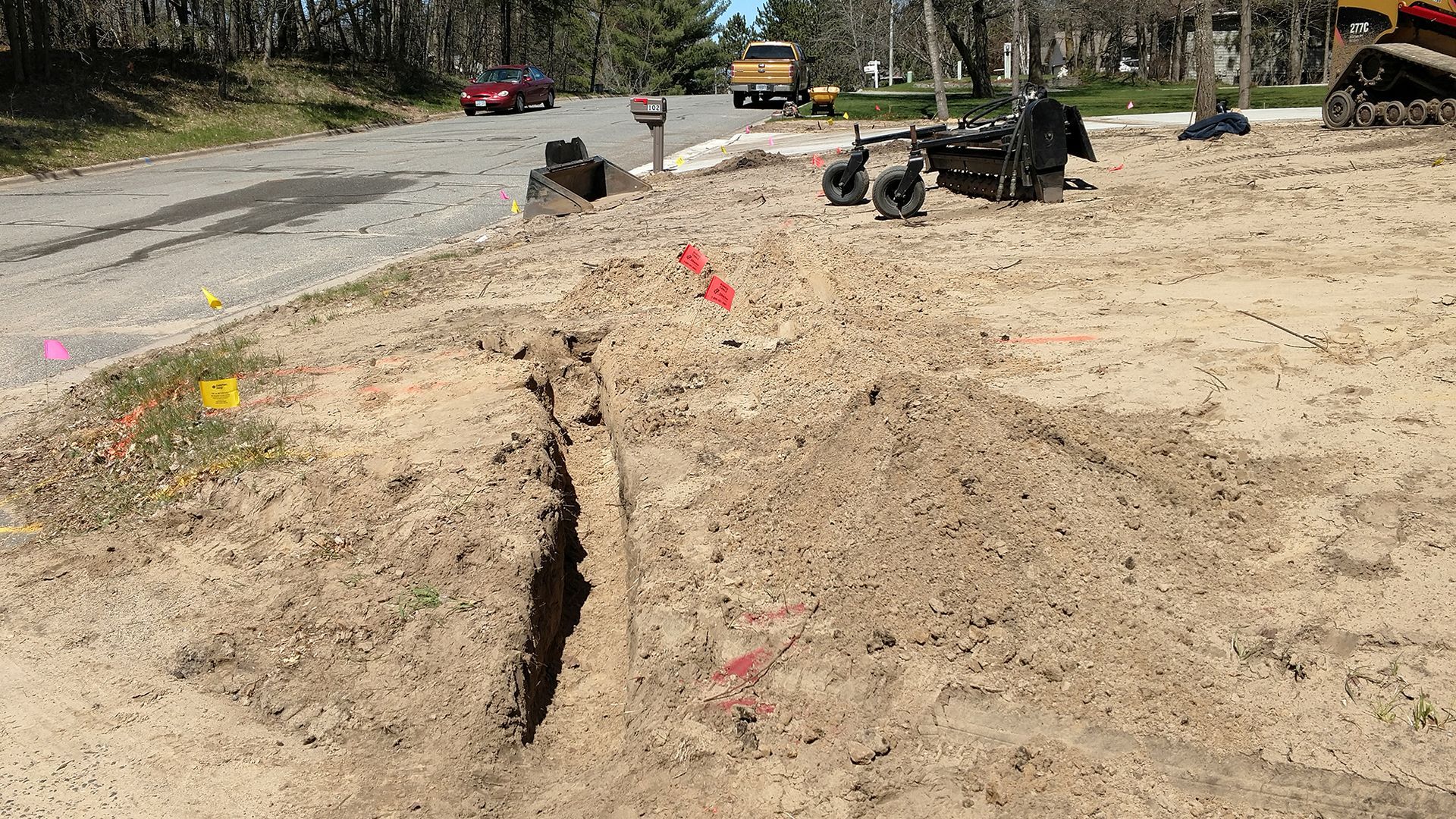 Construction trench in sandy ground beside a dirt road, with heavy equipment in the background.