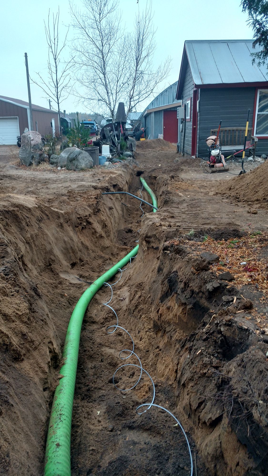 Trench with green pipe running through a muddy yard beside houses and parked vehicles