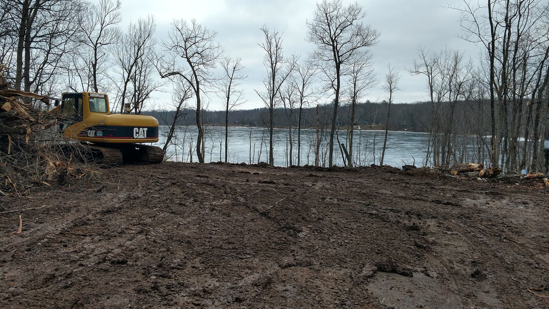 Excavator on a muddy lakeshore with bare trees and calm water under an overcast sky