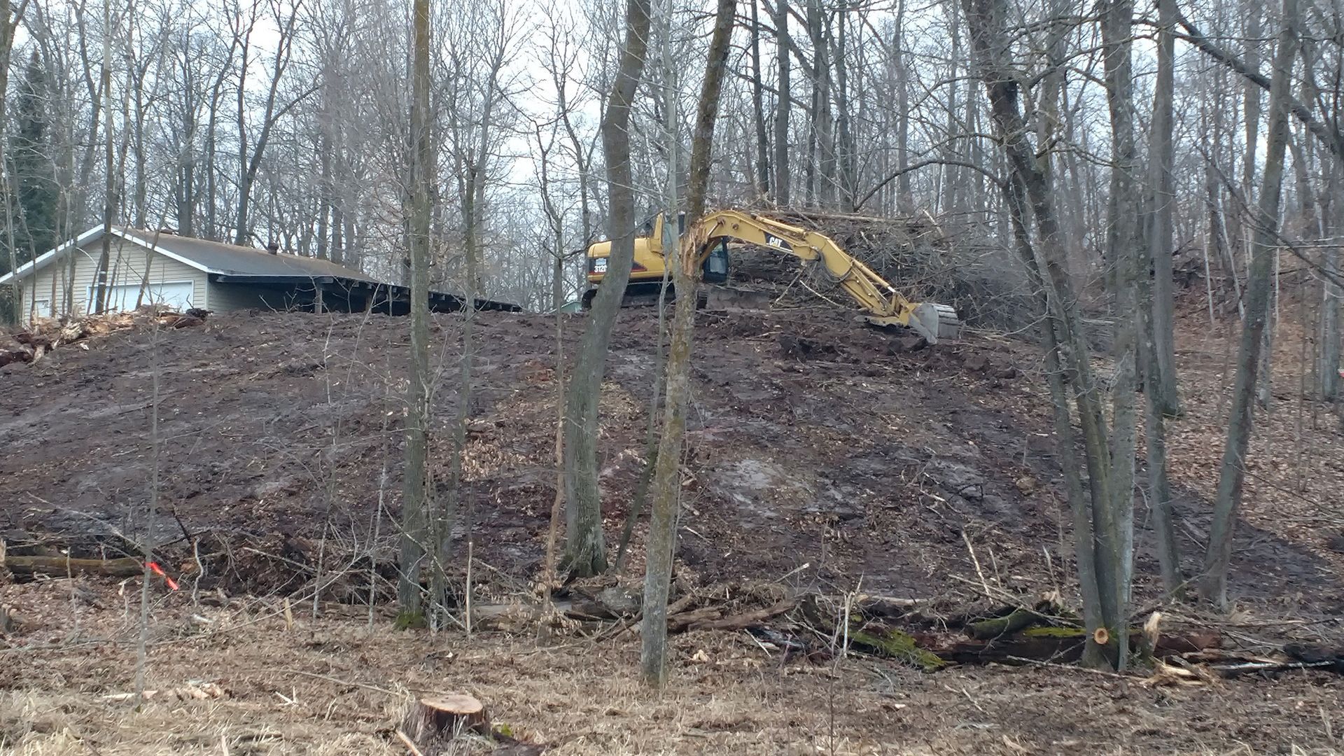 Yellow excavator on a bare wooded hillside near a small house in late fall.