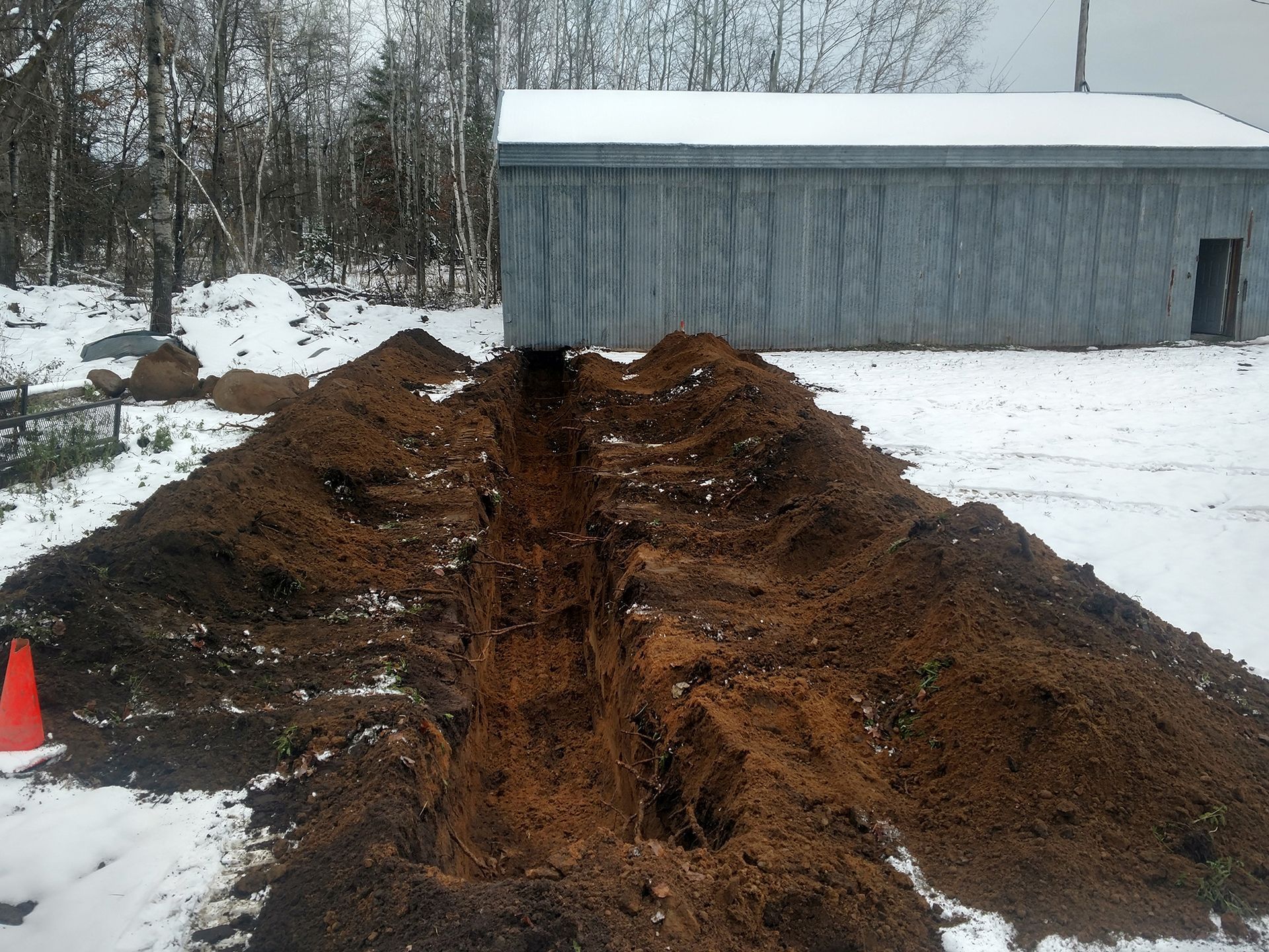 Snowy construction site with a muddy trench leading to a gray shed and orange cone