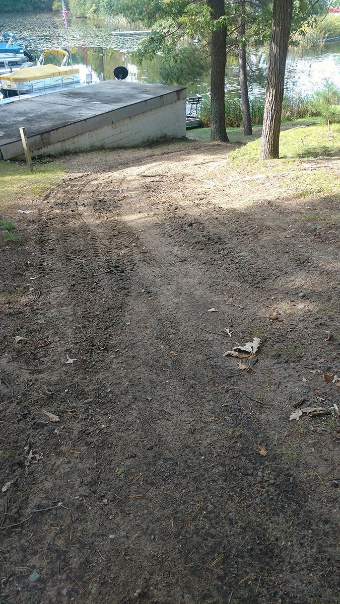 Grassy slope with a dirt path, trees, and a small lakeside dock or boathouse in the background