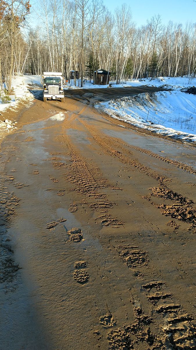 Snowy rural road with muddy tire tracks and a white van near leafless trees.