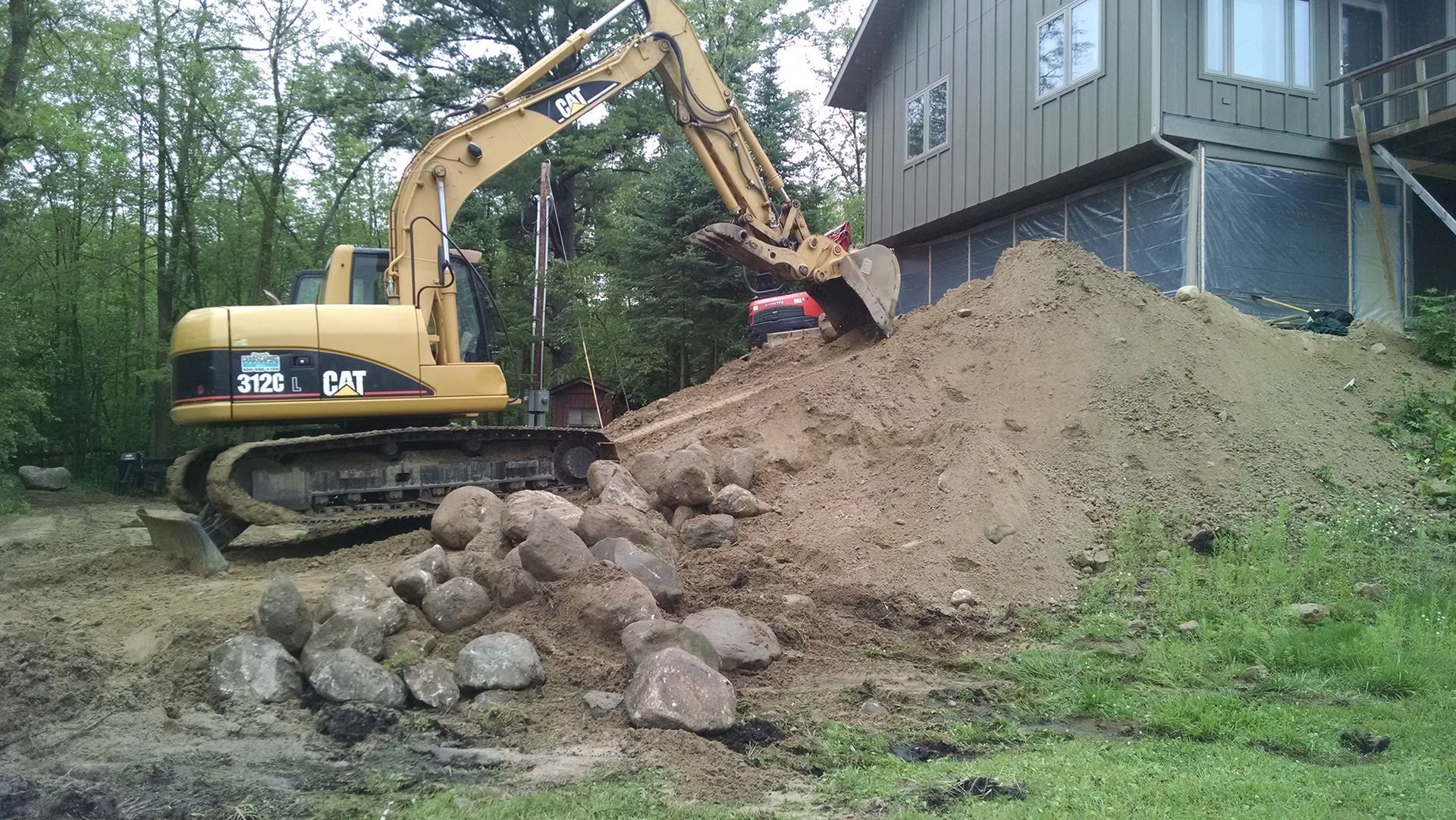 Yellow excavator digging a dirt mound beside a house, with large rocks stacked in the foreground.