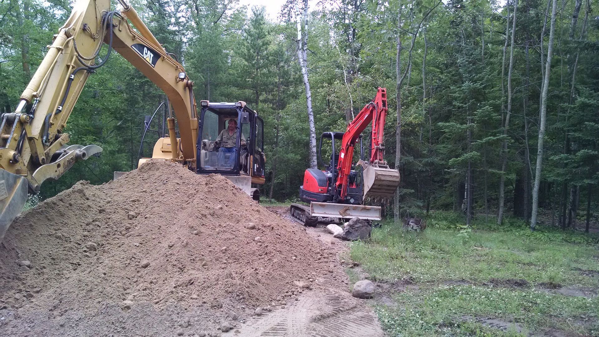 Yellow and red excavators working beside a large dirt pile in a wooded area