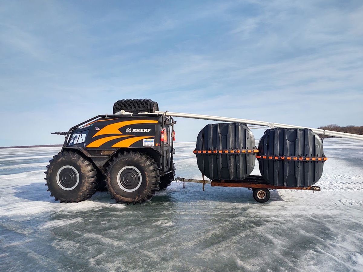 ATV towing a sled loaded with large black barrels across icy terrain under a cloudy sky