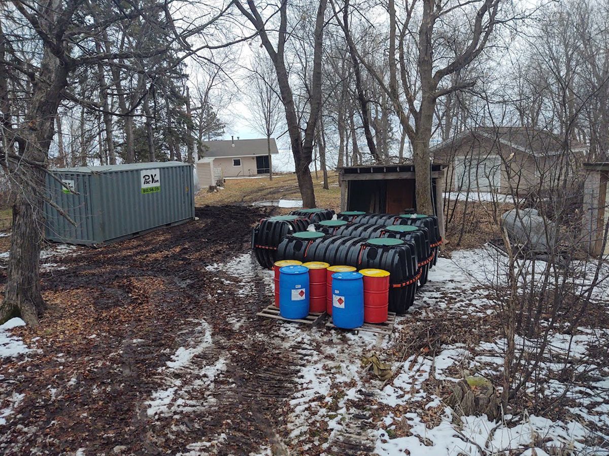 Outdoor yard with stacked colored barrels, a green container, and bare trees in winter