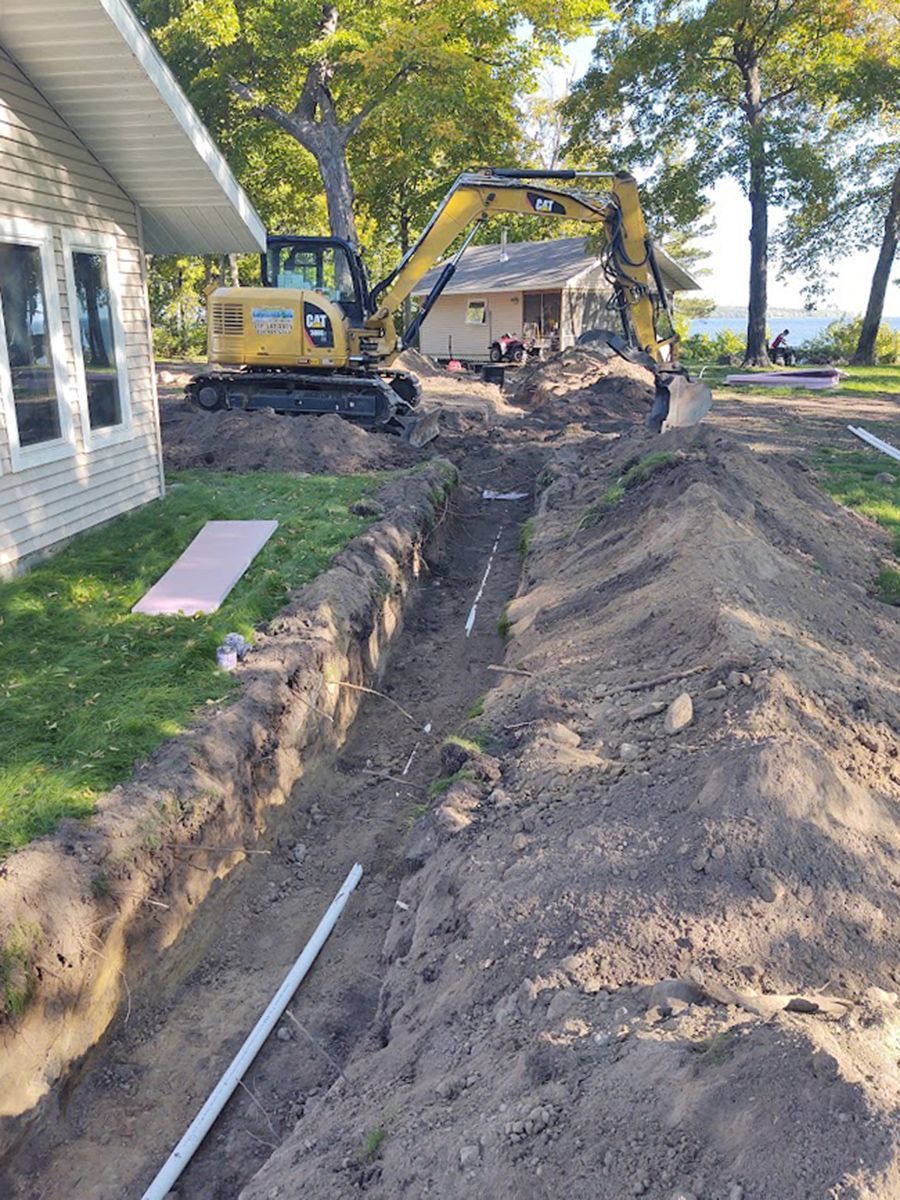 Backhoe digging a long trench beside a house, with dirt piles along the yard.
