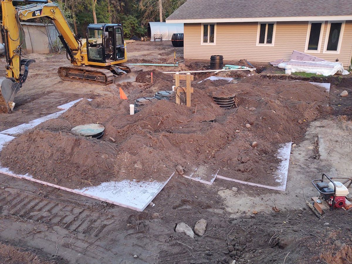Excavator digging a muddy construction site beside a beige house with exposed foundation trenches