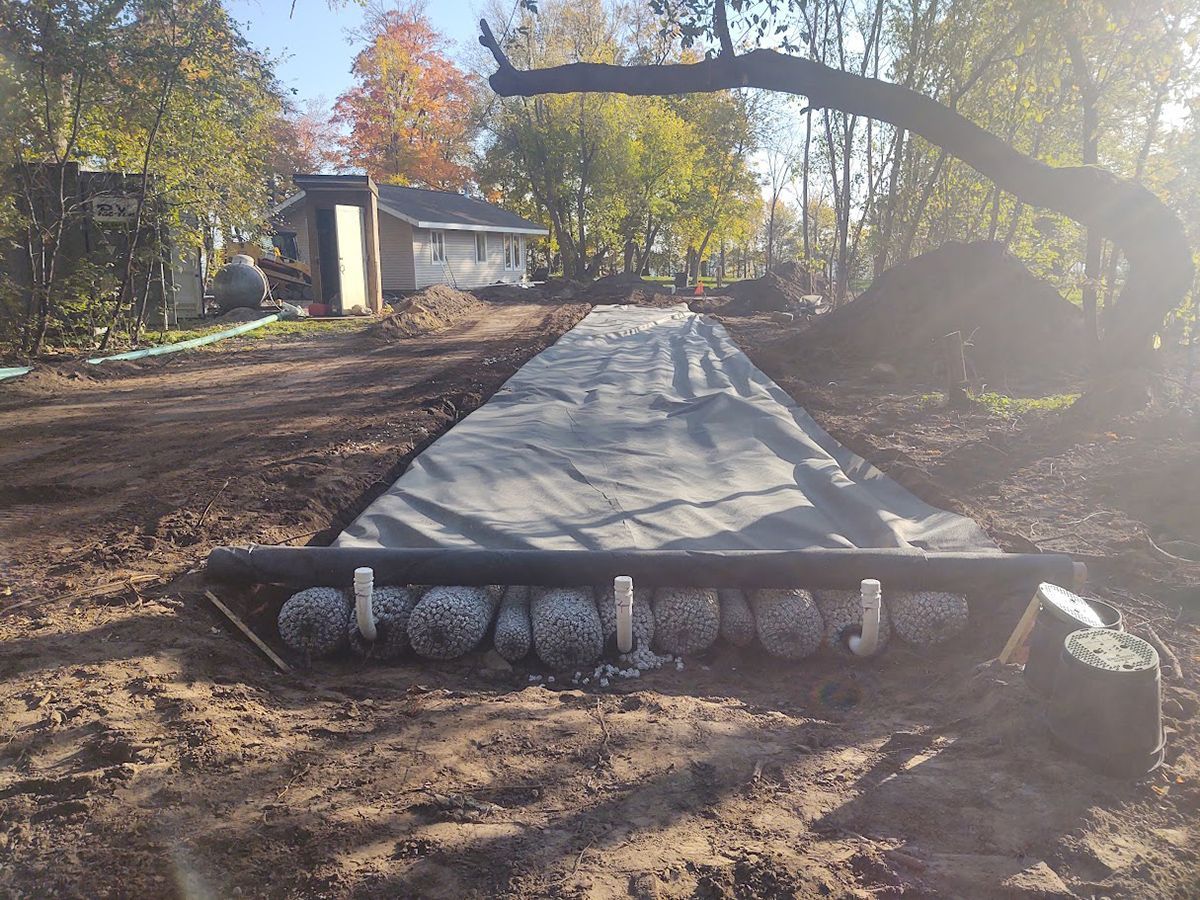 Concrete walkway under construction in a muddy yard, with trees and a small house in the background.