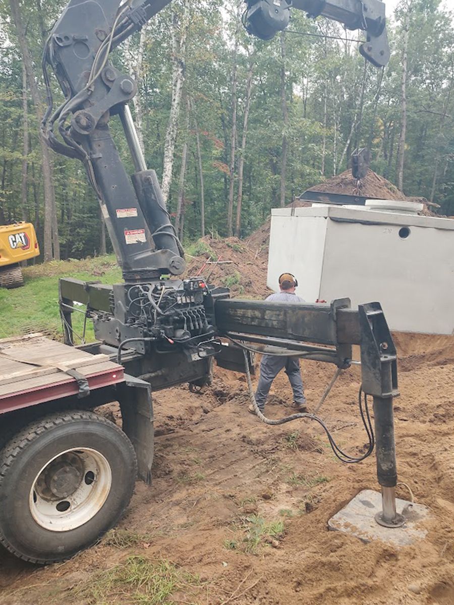 Excavator on a trailer lifting a black crane arm in a wooded worksite, with a white shed in the background