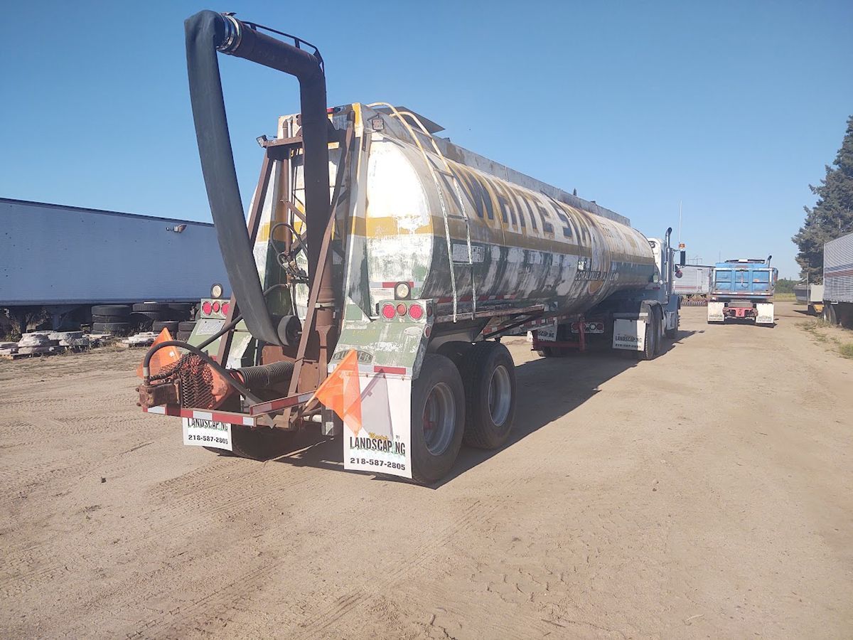 Tanker trailer with red pump equipment parked on a dirt lot under a blue sky, beside another truck.