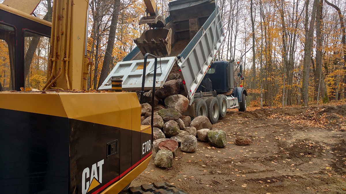Yellow CAT dump truck tipping rocks into a large pile in a wooded work site.