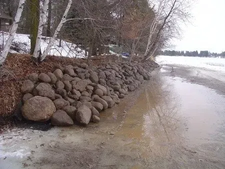 Rocky shoreline with piled boulders beside a snowy, muddy riverbank and calm water