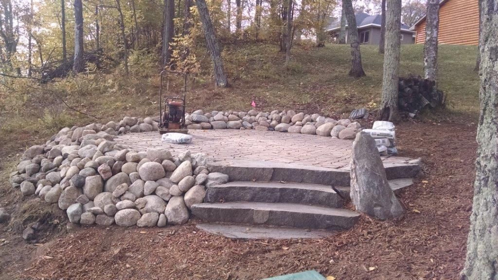 Stone steps leading to a raised circular rock patio in a wooded yard, with a small sculpture on top.
