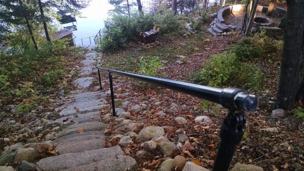 Stone steps descending through a wooded lakeside path with a black handrail and autumn leaves.