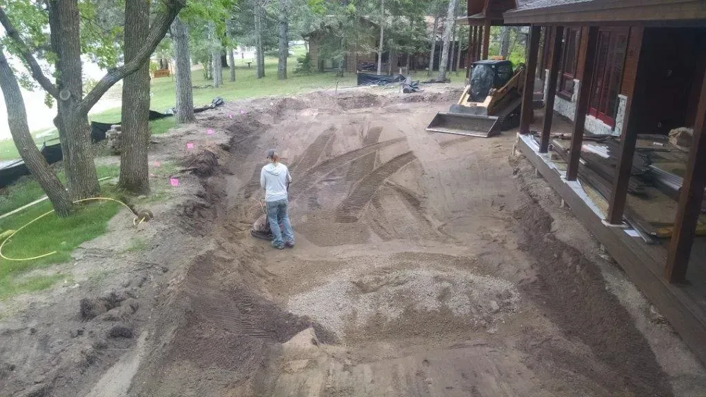 Worker grading a dirt yard beside a house, with trees and a porch in the background.