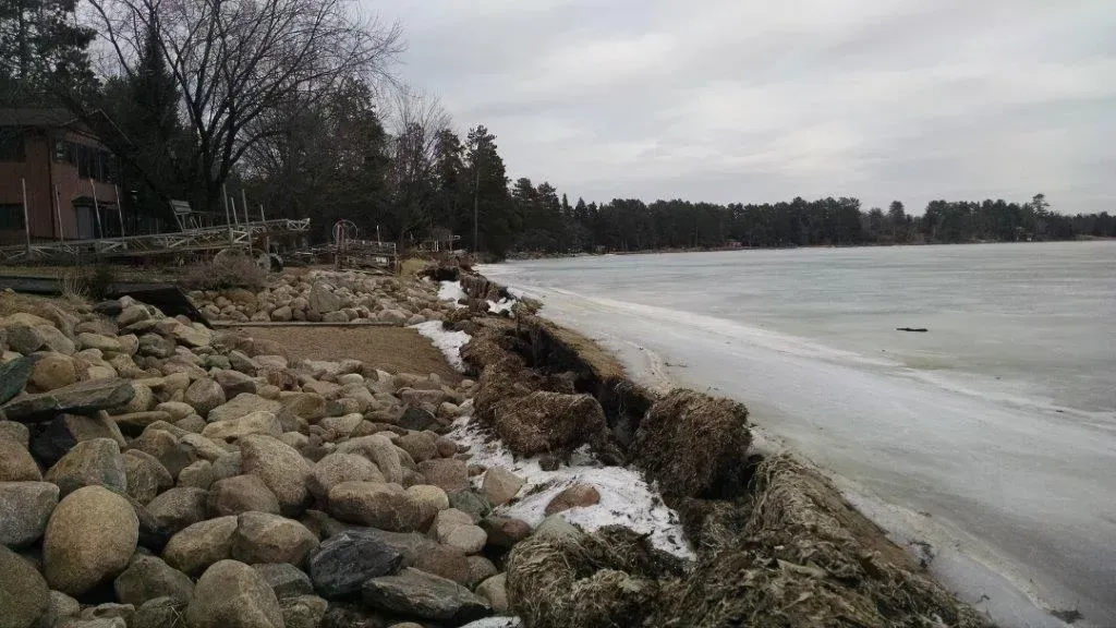Rocky shoreline beside a frozen lake under a cloudy sky, with houses and trees along the coast