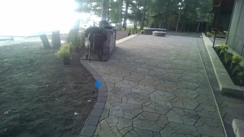 A stone walkway beside the water with a seated person near the path and trees along the edge.