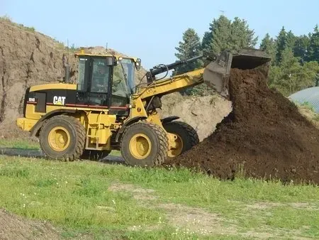 Yellow CAT loader dumps dirt into a large pile in a grassy outdoor worksite.
