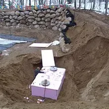Pink box-shaped object on a dirt slope with a plank ramp, near a stone retaining wall and snow patches