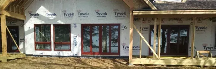 House under construction with red-trimmed windows and doors, framed walls, and exposed roof sheathing