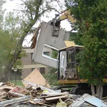 Collapsed house being lifted by an excavator, with debris scattered in front and trees nearby
