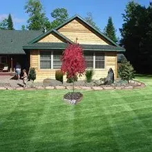 A house with a green lawn, a red tree in front, and trees and rocks in the landscaped yard.