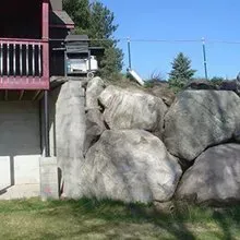 Person climbing over large boulders beside a house with a red deck and blue sky