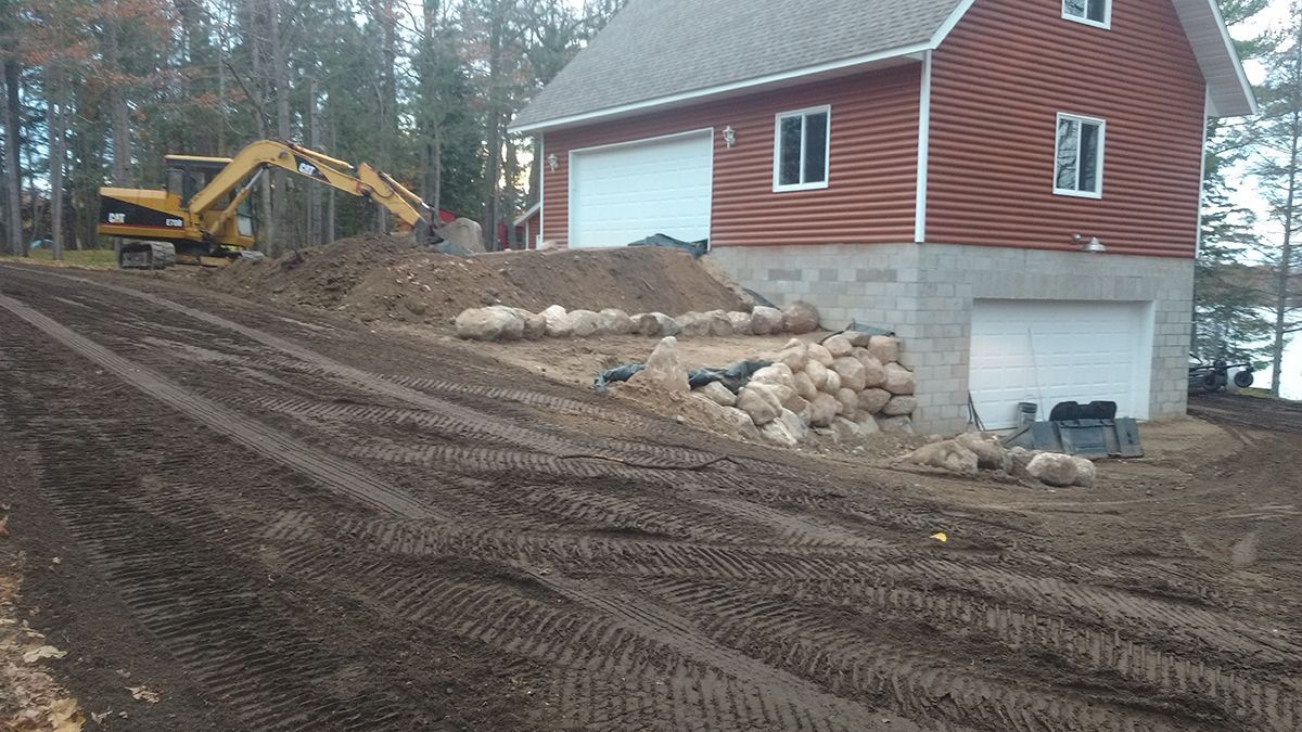 Mud-covered construction site beside a red house with a yellow excavator and dirt driveway.