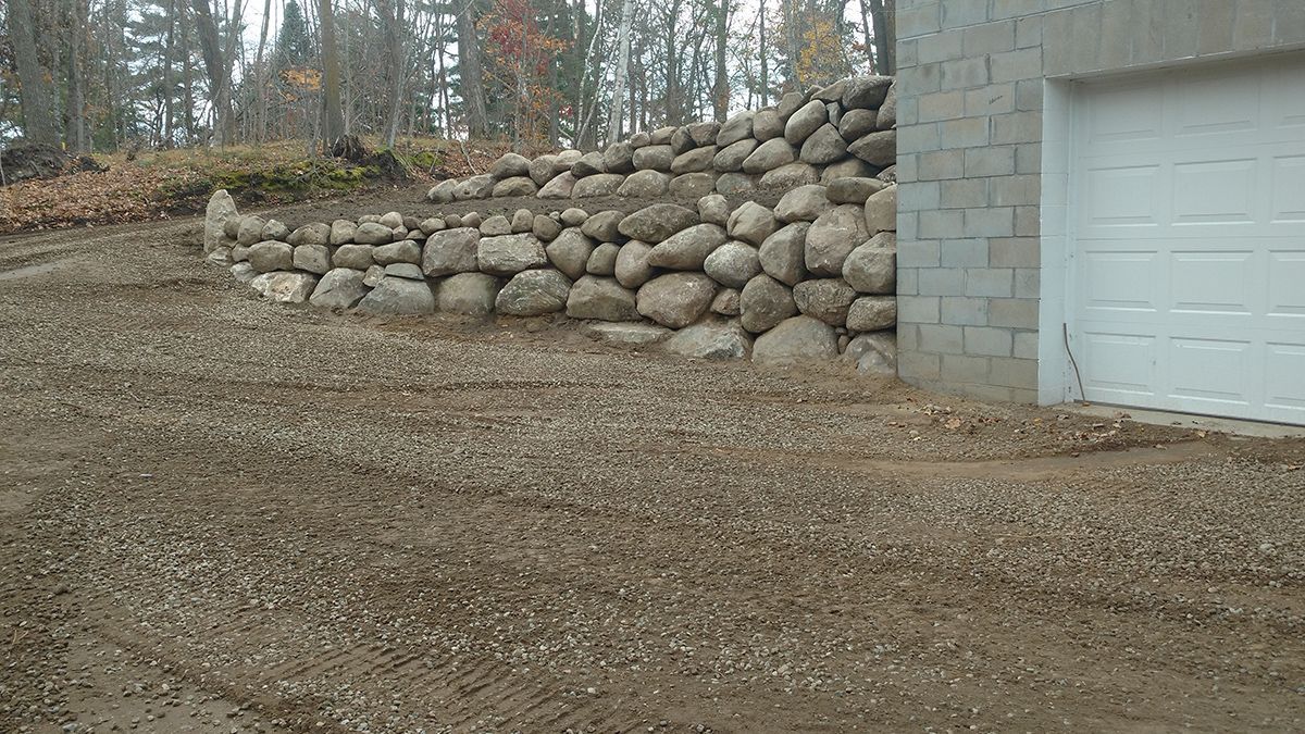 Rock retaining wall beside a white garage door on a gravel driveway in a wooded area