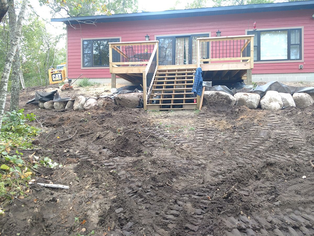 Red house with wooden porch and stairs above a muddy, cleared yard surrounded by rocks and trees