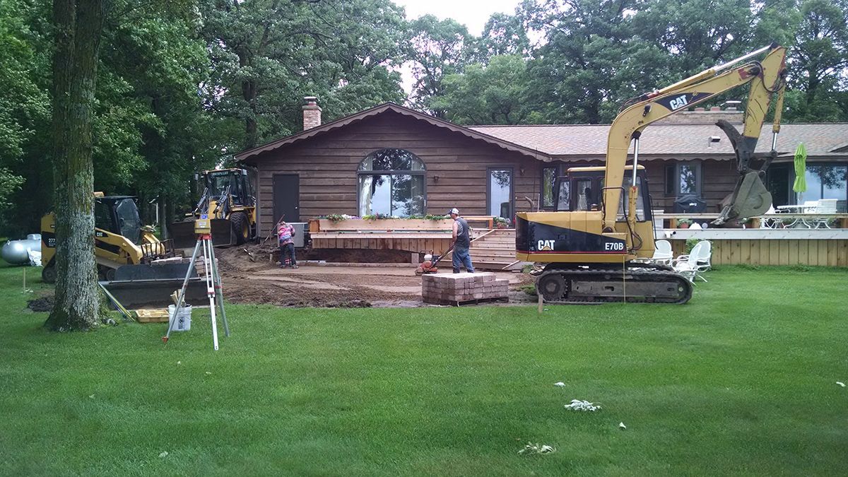 Excavator dismantling a brick house in a grassy yard with a person standing nearby.